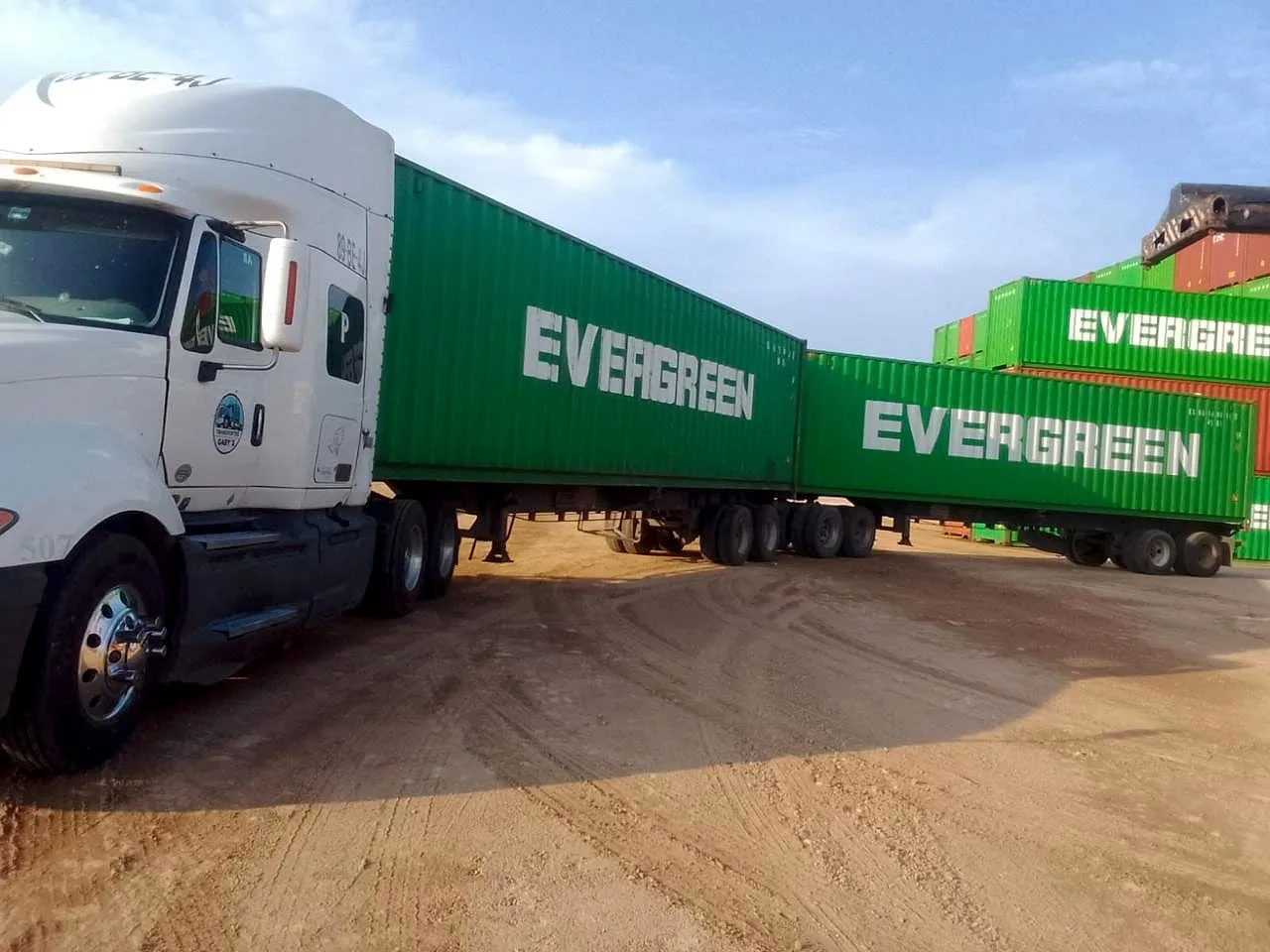 Tractocamión blanco número 507 con dos contenedores verdes en un patio de tierra.