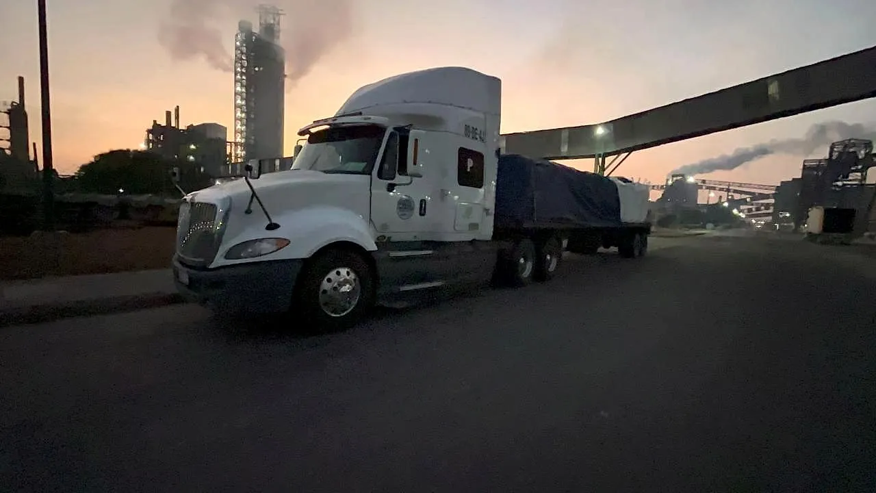 Tractocamión blanco con plataforma encarpada frente a una planta industrial al anochecer.